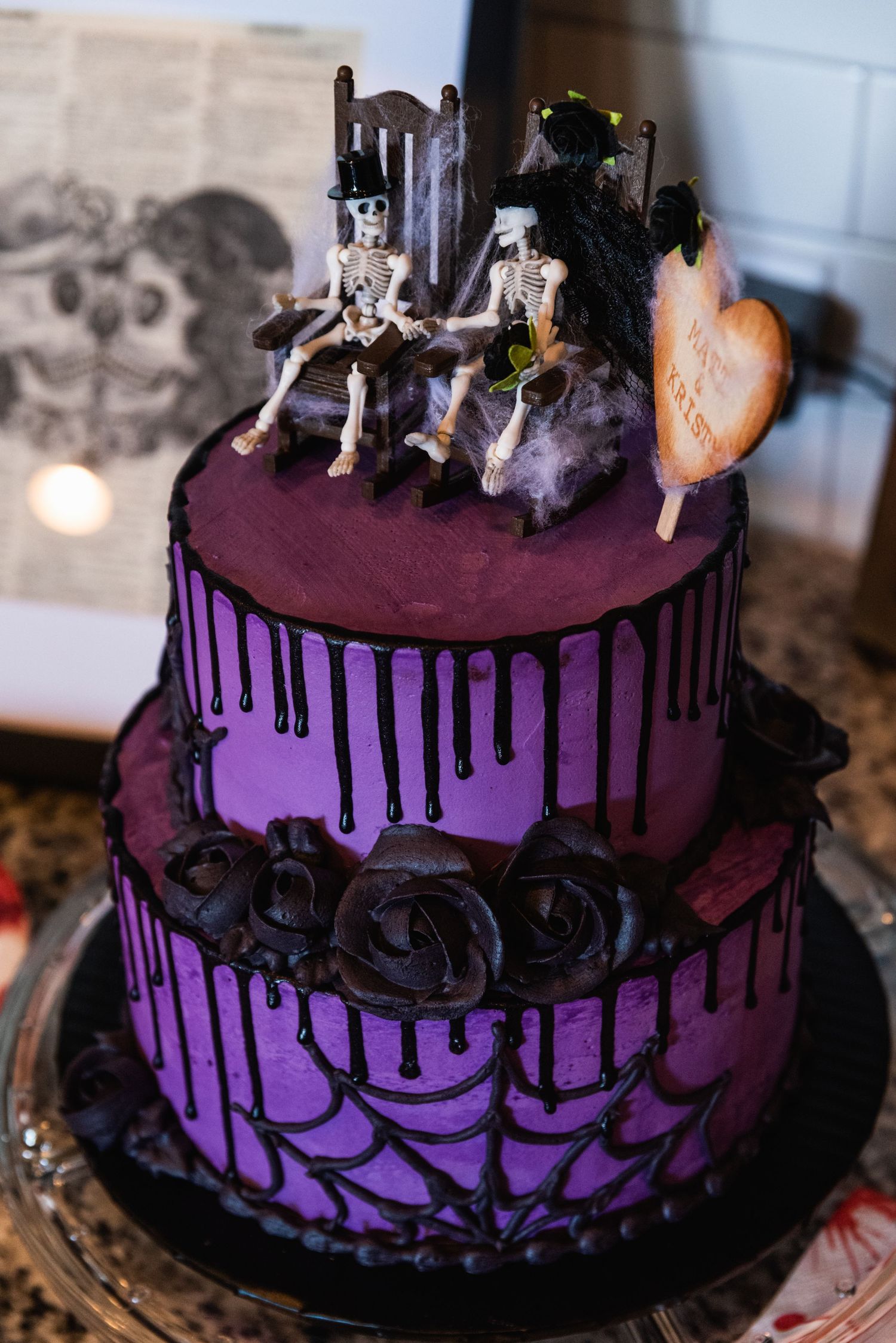 A purple tiered wedding cake decorated with black roses and skeleton figurines on top.
