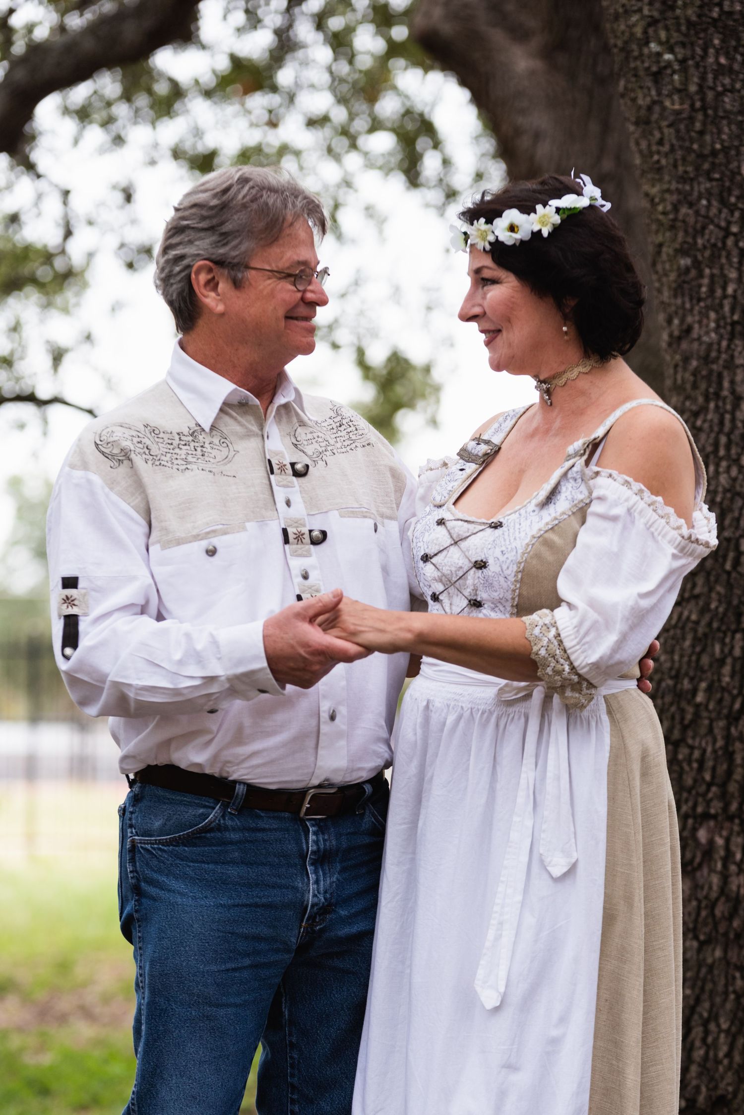 A couple shares an intimate moment while holding hands outdoors under tree branches.