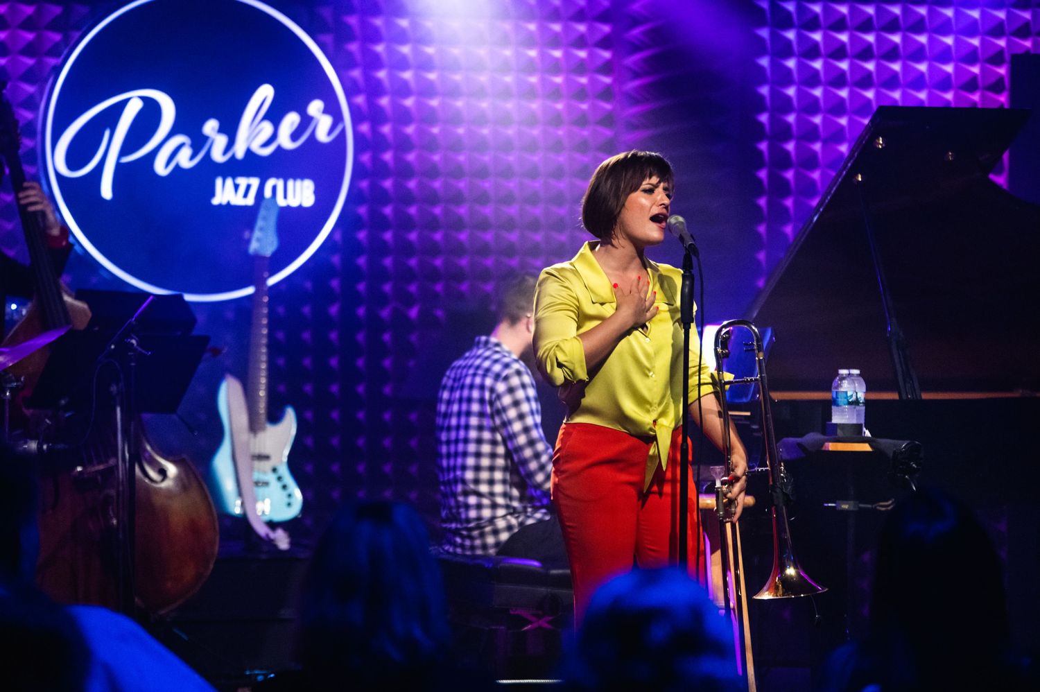 A performer in yellow and red sings on stage at Parker Jazz Club with vibrant purple lighting.