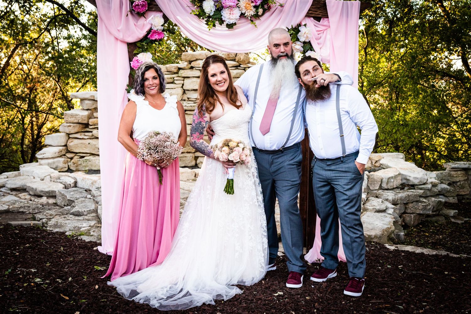 Wedding party posing under floral arch with stone wall backdrop and pink decor.