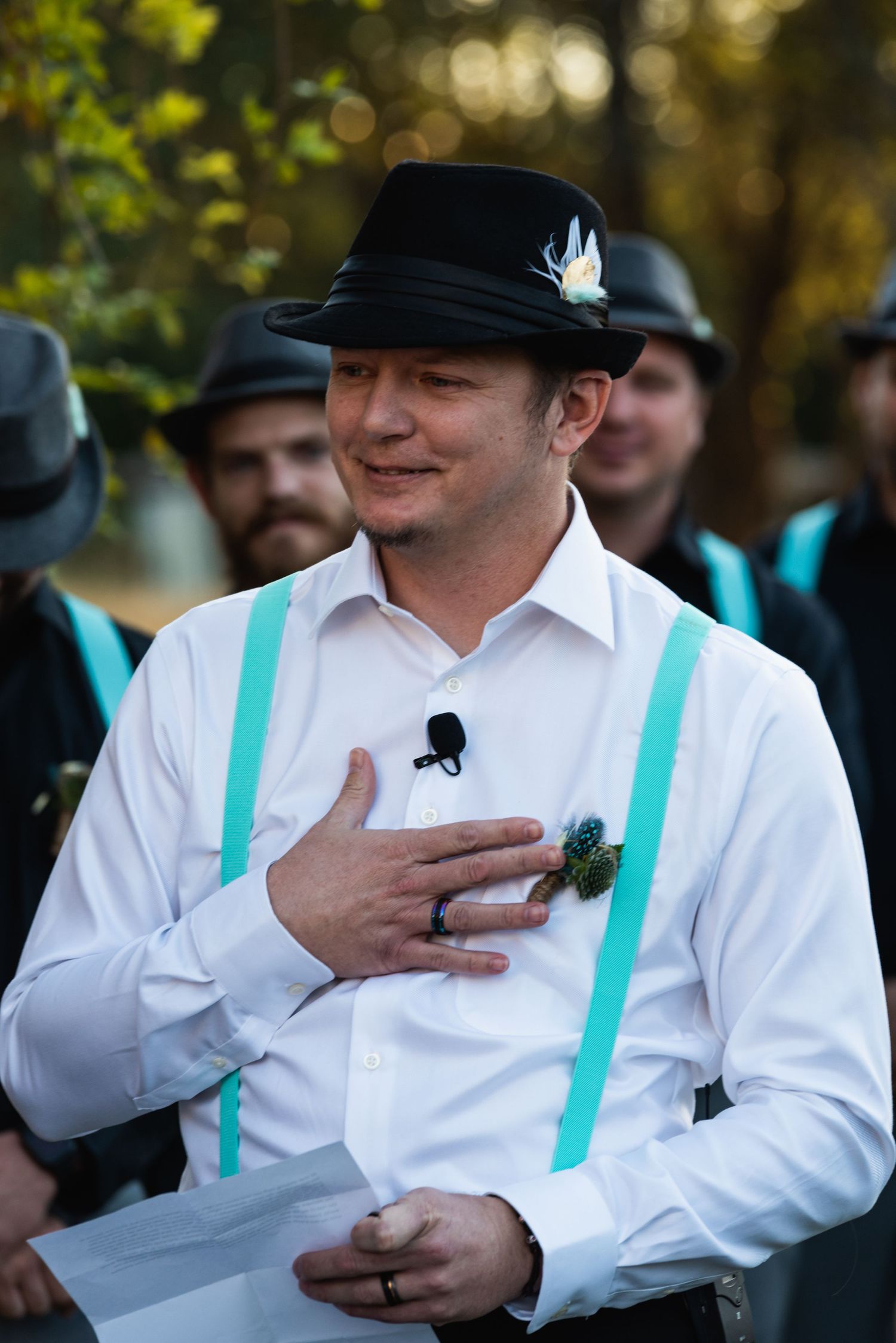 A groom wearing turquoise suspenders and a black hat touches his heart during the ceremony.
