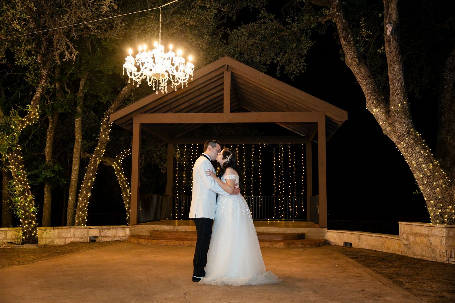 Bride and groom share a romantic moment under a grand chandelier at an elegant outdoor evening wedding venue.