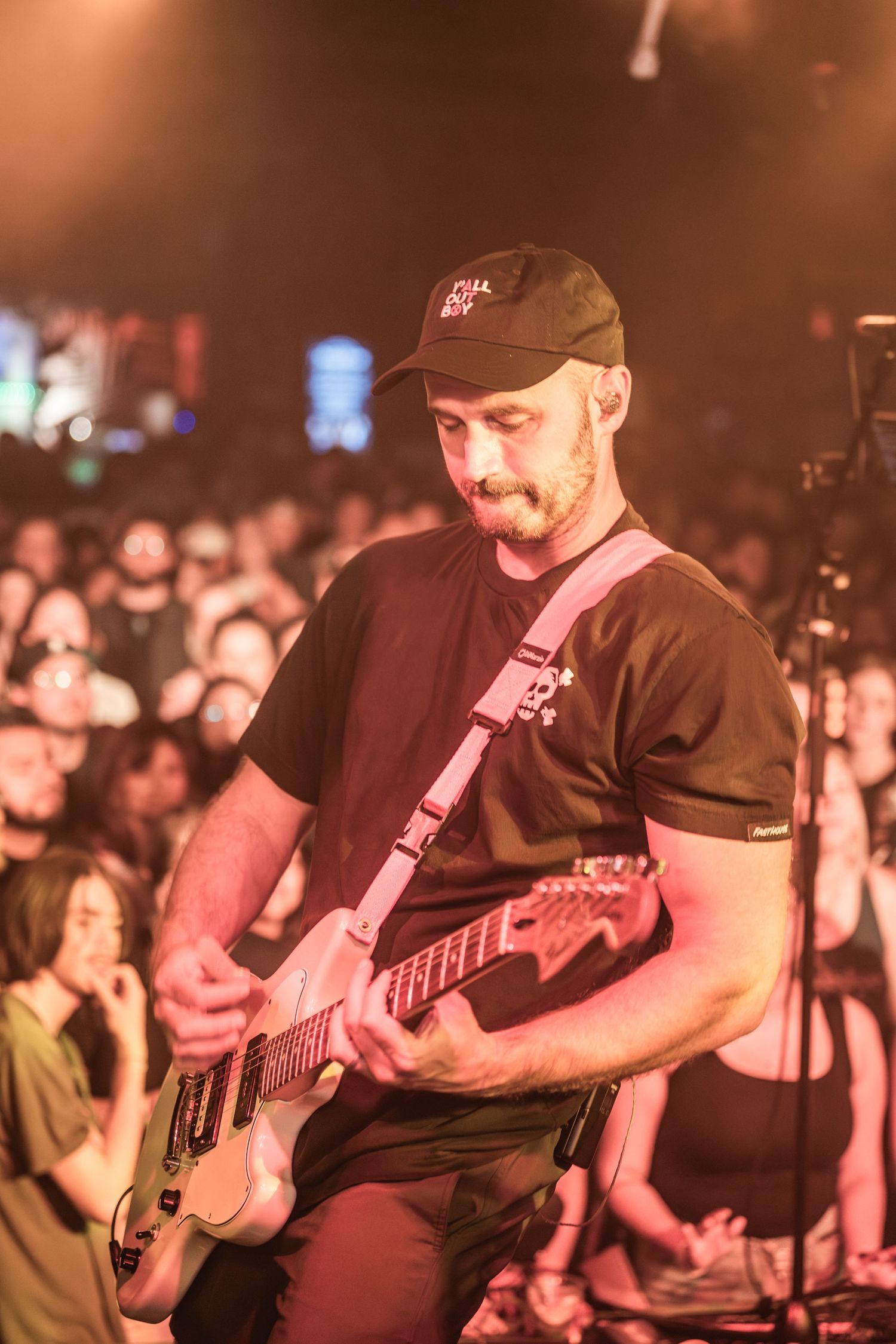 A guitarist performs on stage in a black t-shirt and cap while playing an electric guitar in front of a crowded venue.