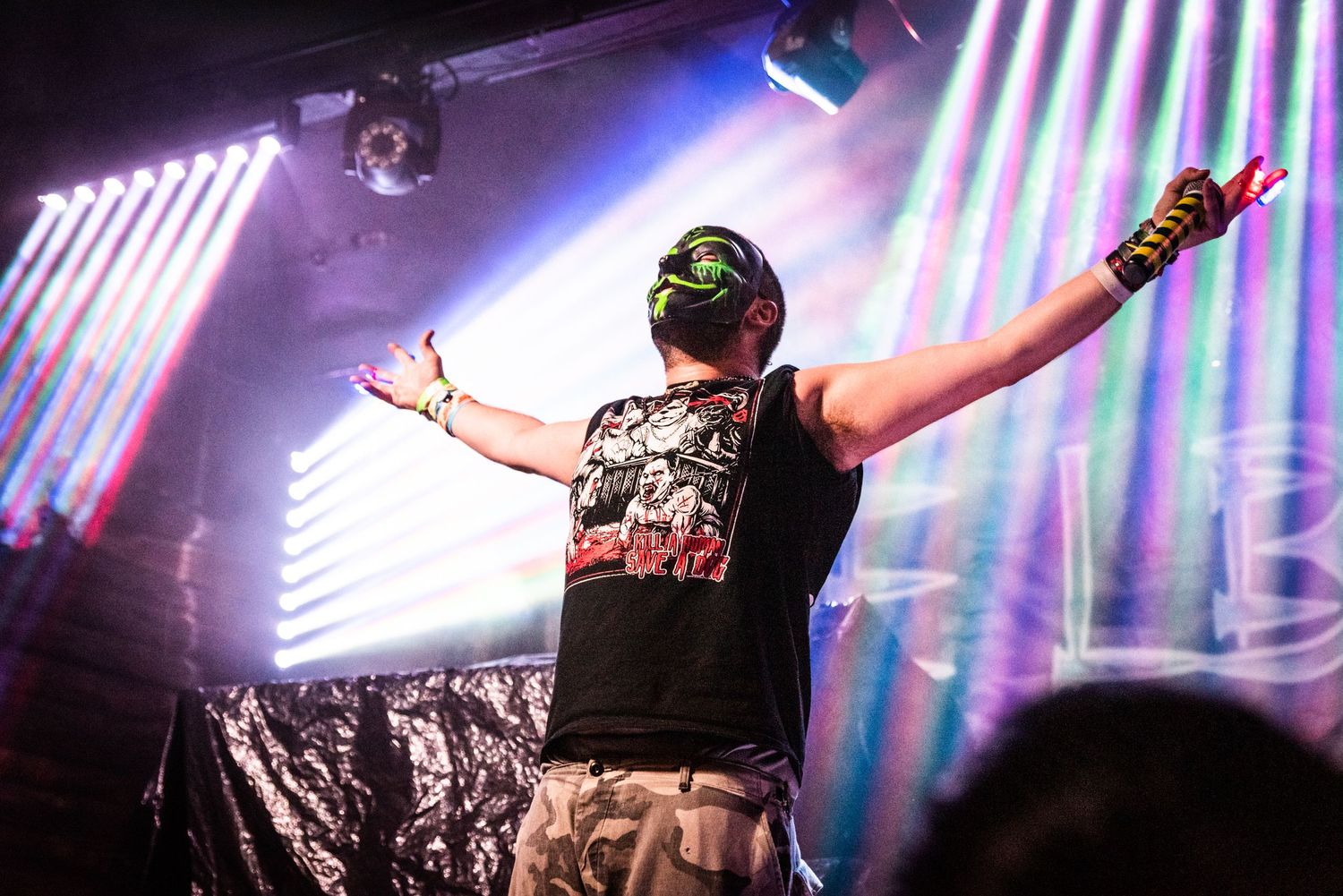 Concert performer with raised arms silhouetted against colorful striped lighting backdrop.