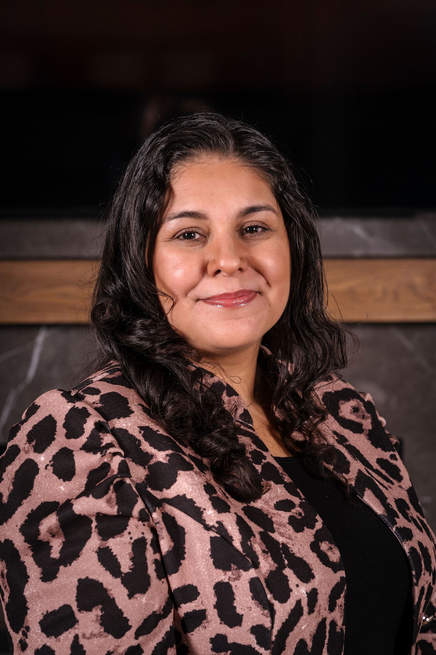 A professional portrait of someone wearing a pink and black patterned blouse against a dark background.