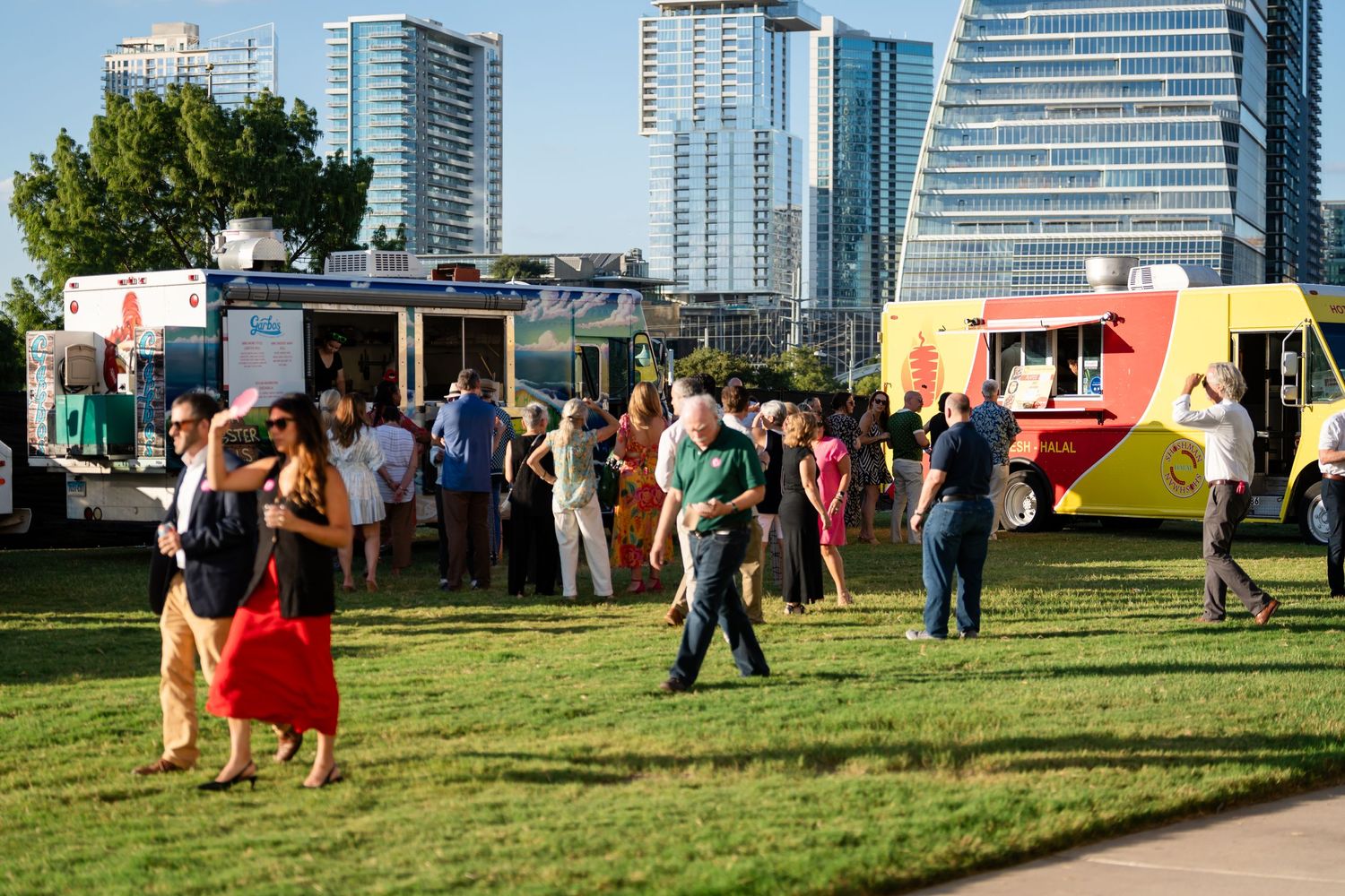 Crowd gathered at food truck festival on grass field with modern city skyline in background.