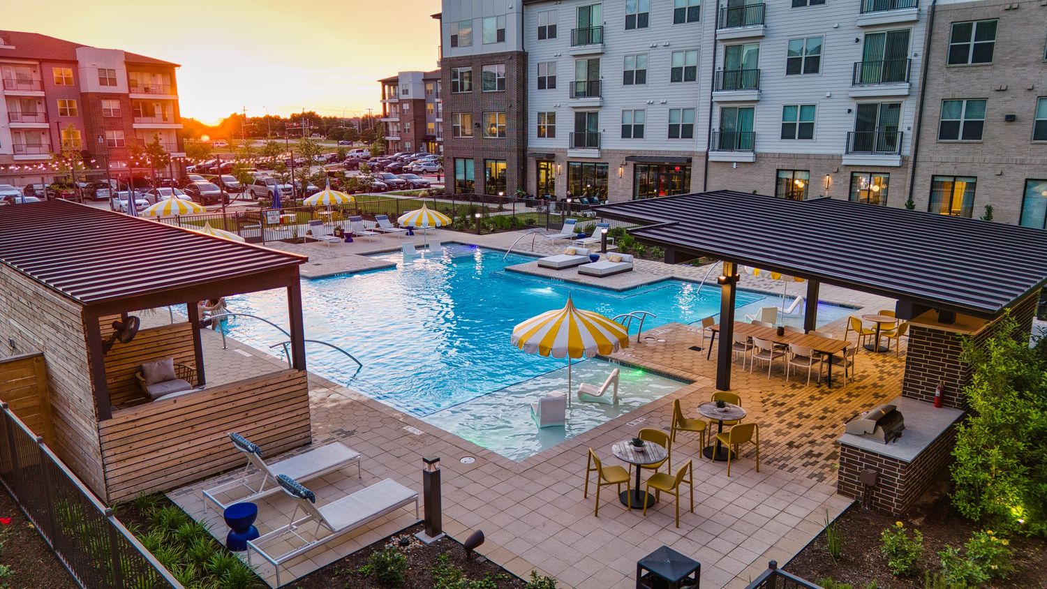 Sunset view of apartment complex pool area with wooden cabanas and yellow umbrellas.