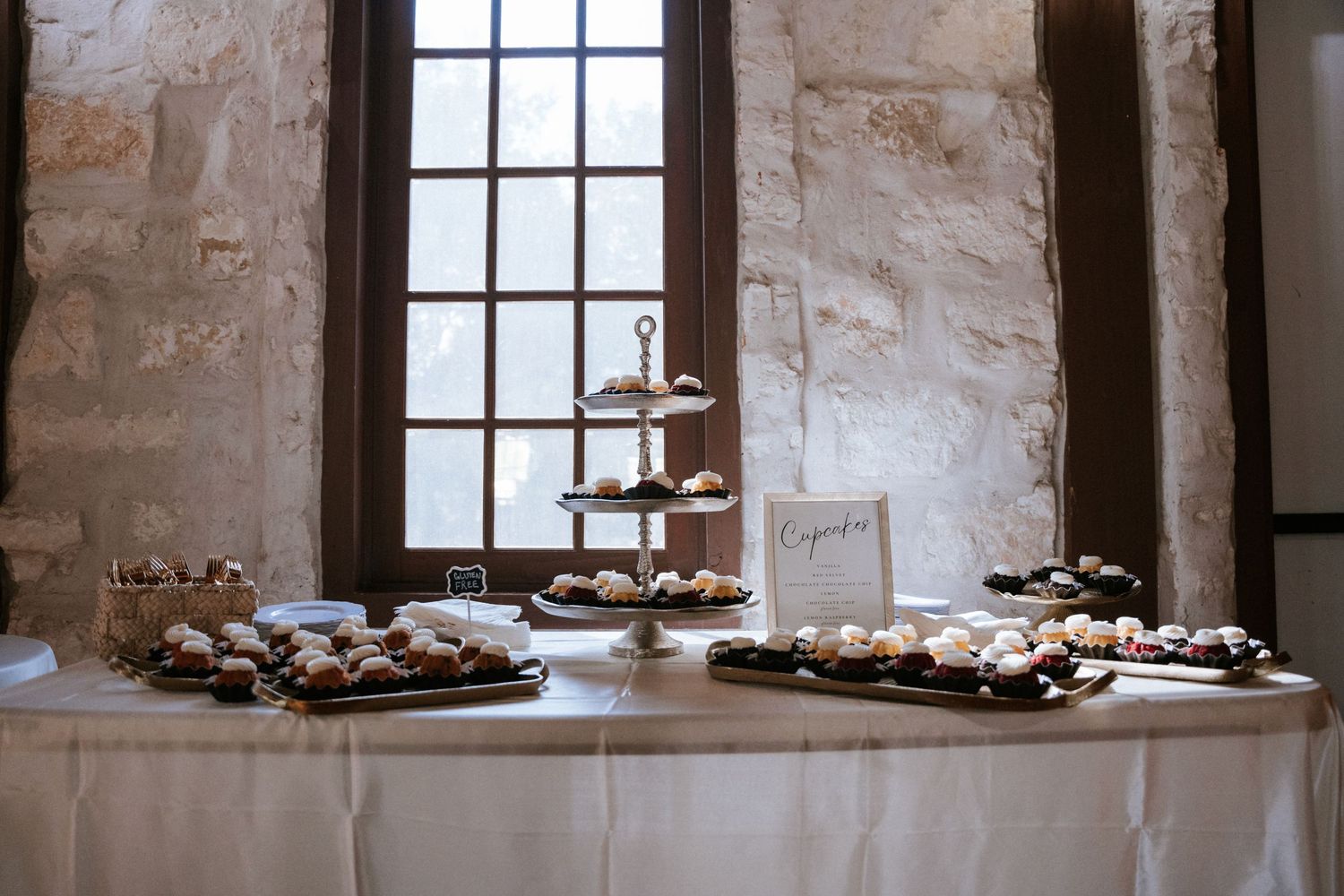 An elegant dessert table display with cupcakes and pastries arranged on white tablecloths near a window.