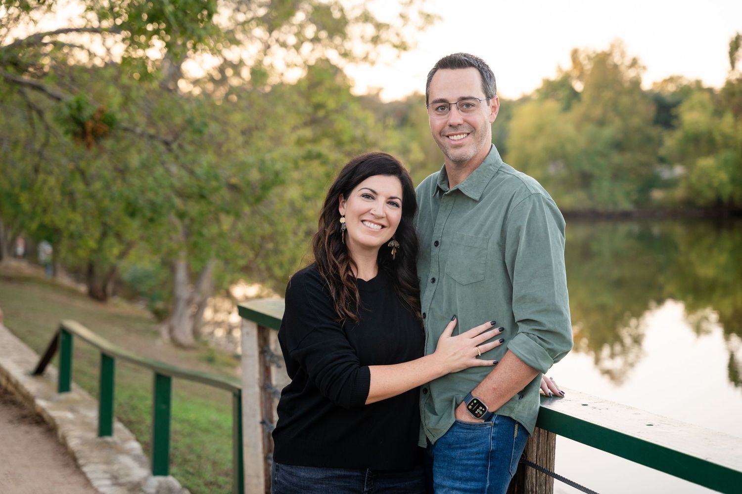 A couple poses together on a wooden bridge with a serene pond and autumn trees in the background.