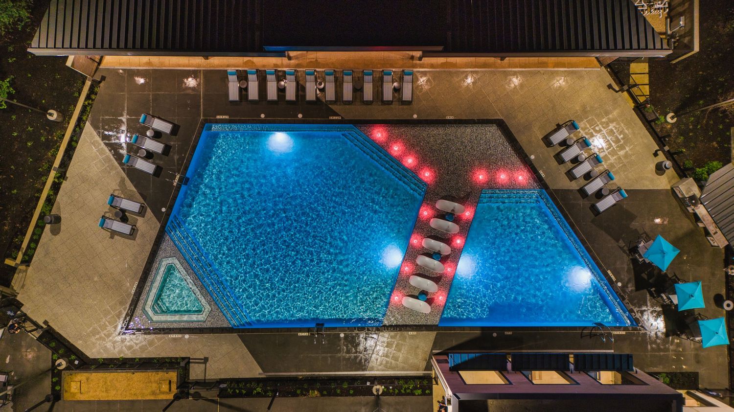 Aerial view of a geometric swimming pool with red loungers and blue lighting at night.