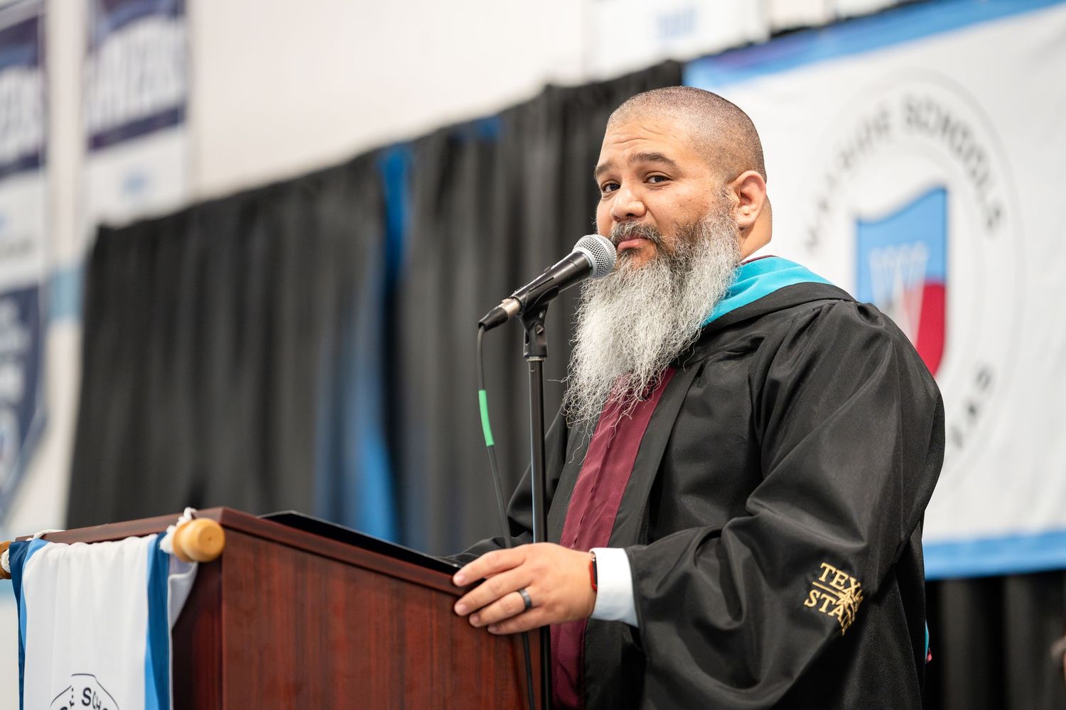 Speaker with grey beard addresses audience from podium at graduation ceremony.