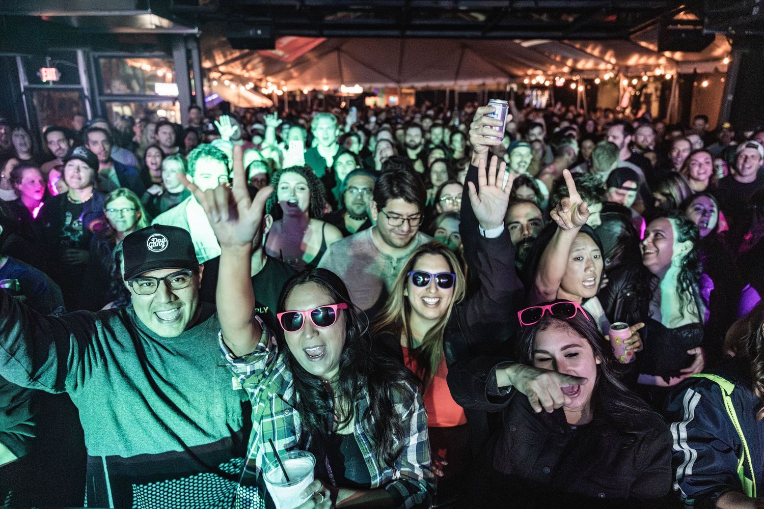 Energetic crowd at a nightclub illuminated by colorful lights with their hands raised during a live music performance.