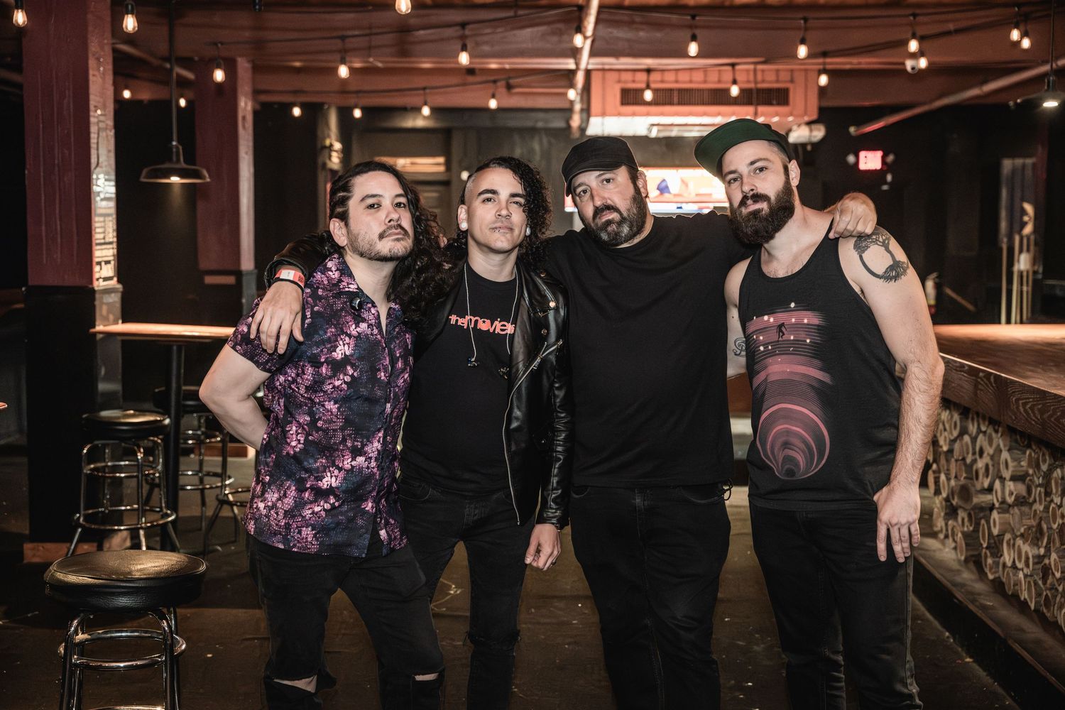 Four band members pose together in casual attire at bar with string lights overhead.