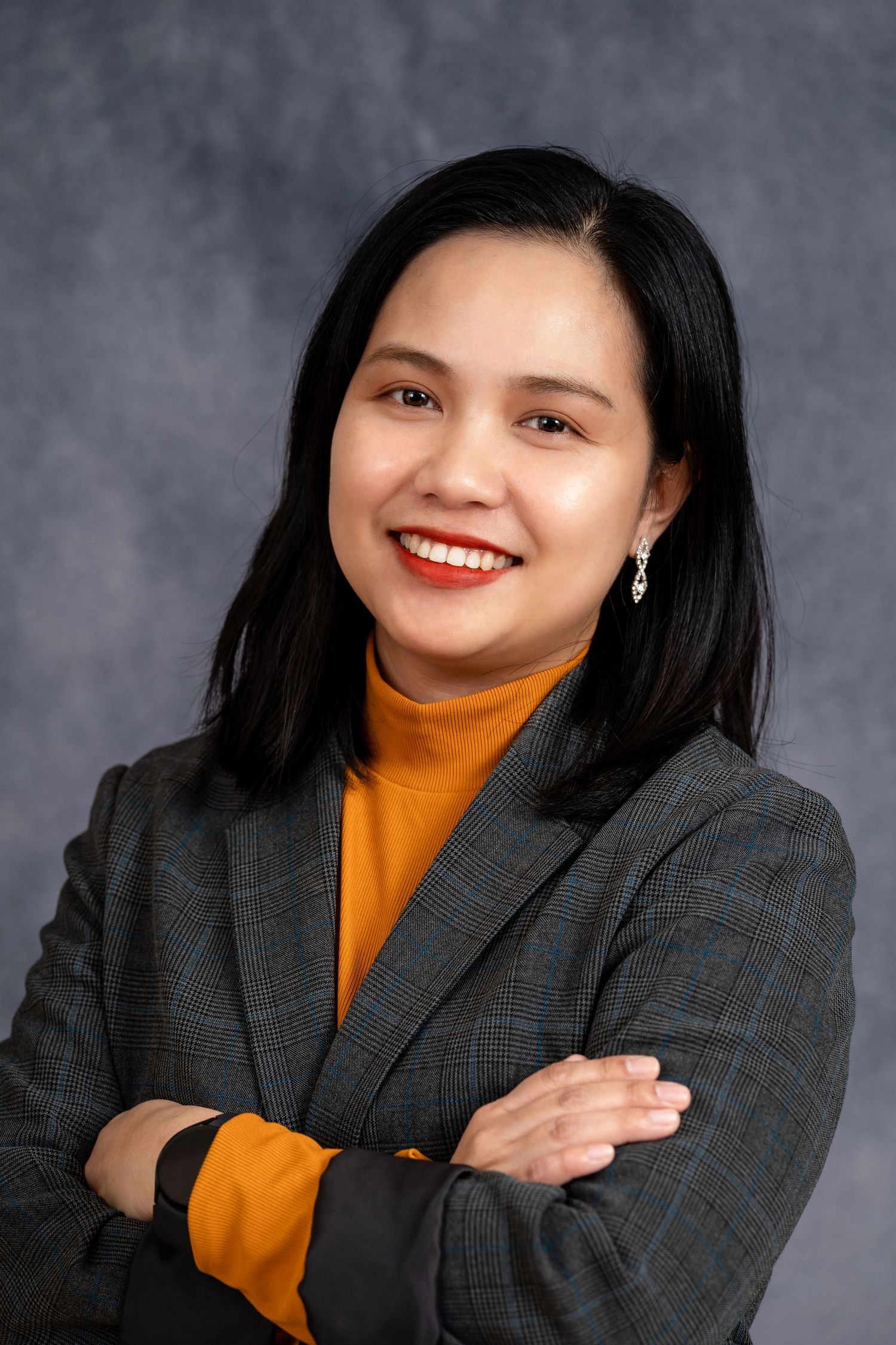 Professional headshot of a person in an orange turtleneck and plaid blazer against a gray background.