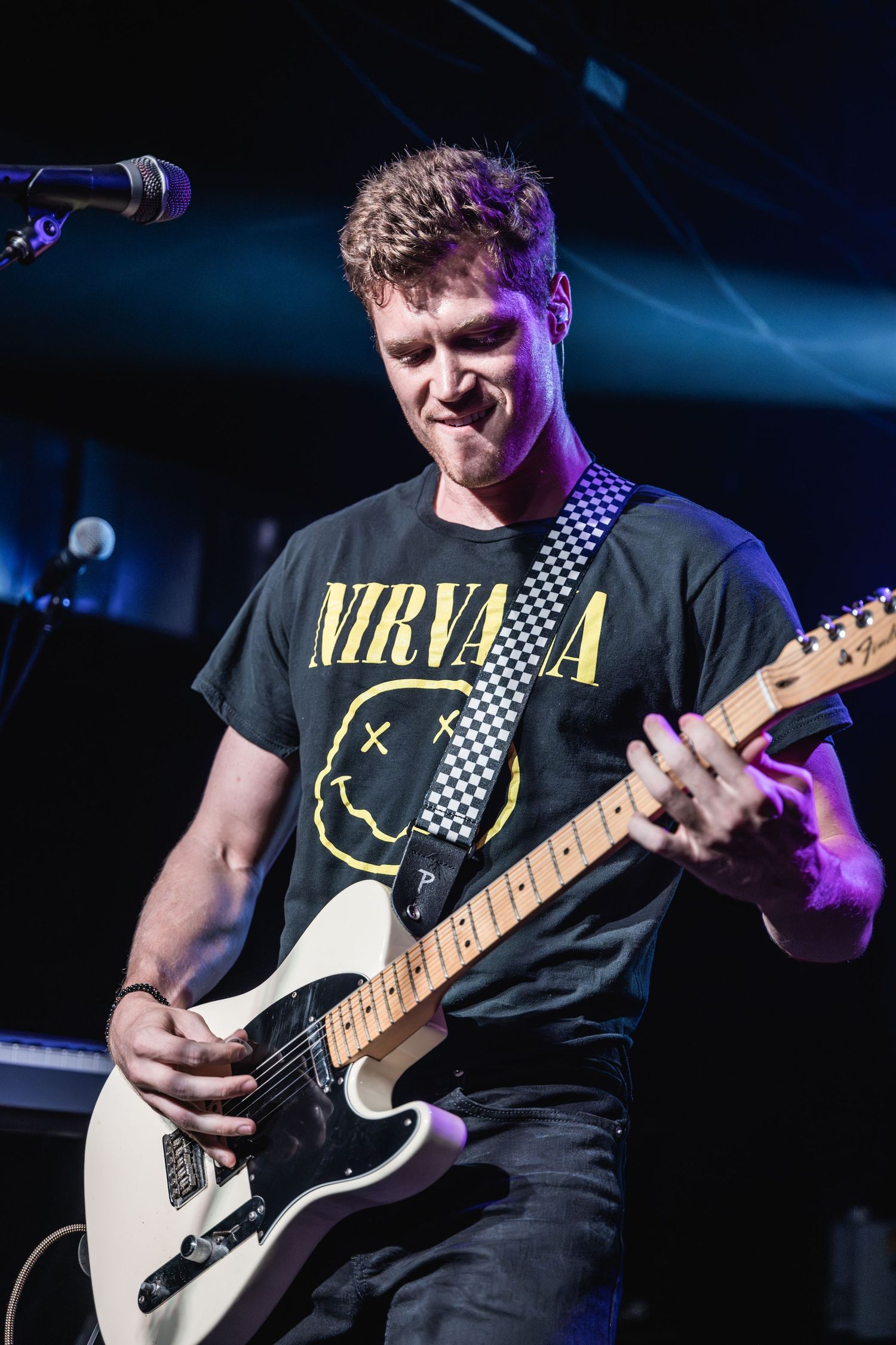 Musician plays electric guitar while wearing a Nirvana t-shirt under blue and purple stage lighting.
