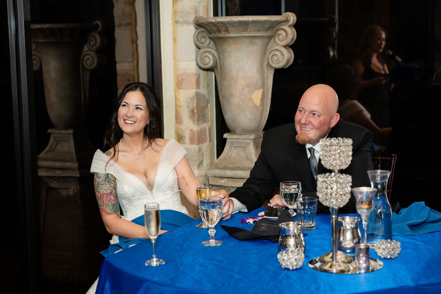 Couple seated at head table with elegant stone columns and blue tablecloth.