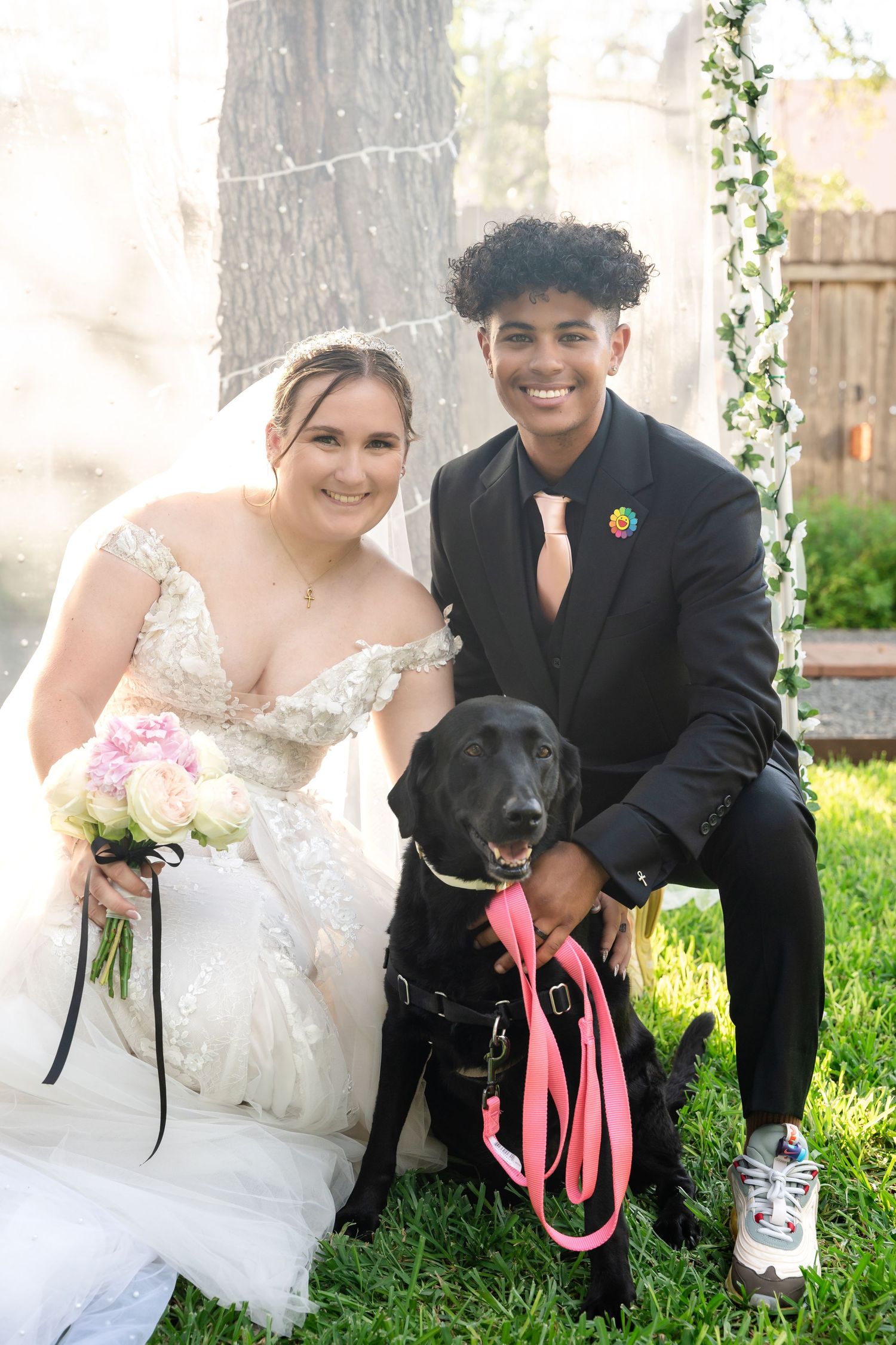 Newlyweds pose with their black dog wearing a pink leash under a floral wedding arch.