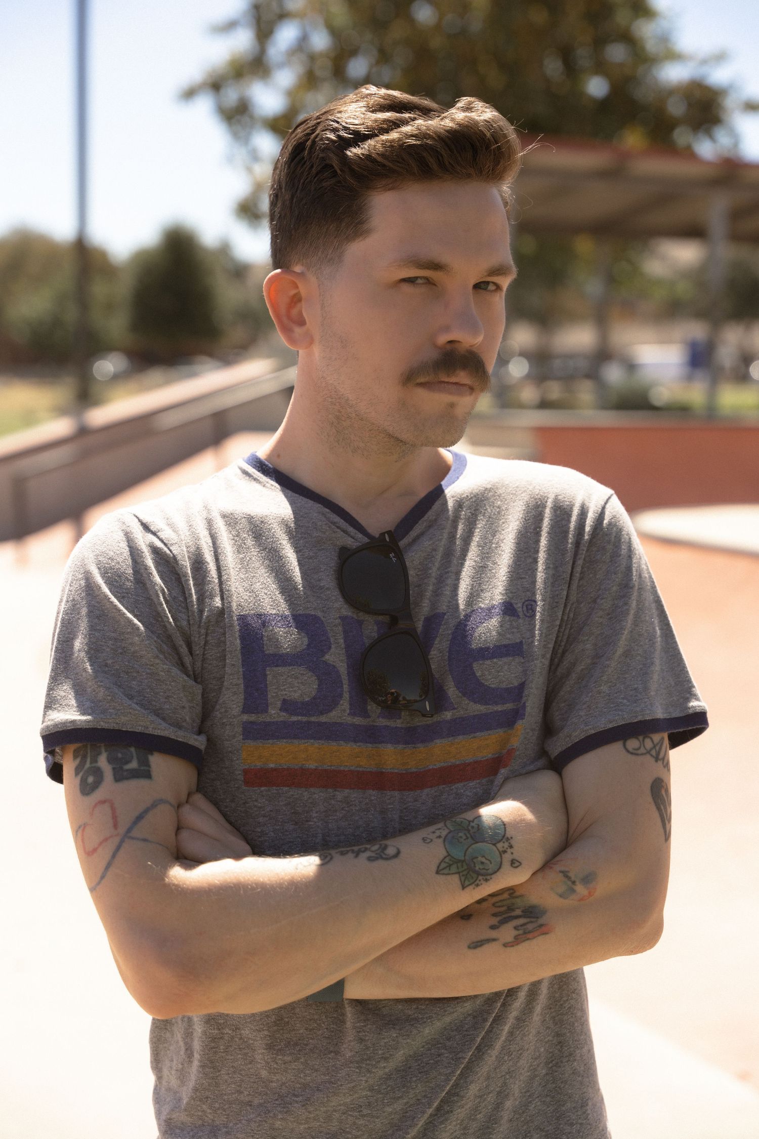 A person in a gray BICE t-shirt with visible tattoos stands with arms crossed outdoors.