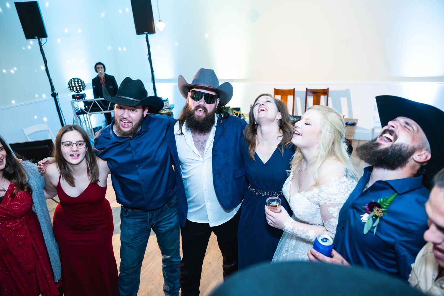 Group of party guests laughing together at a wedding reception wearing cowboy hats and formal attire.
