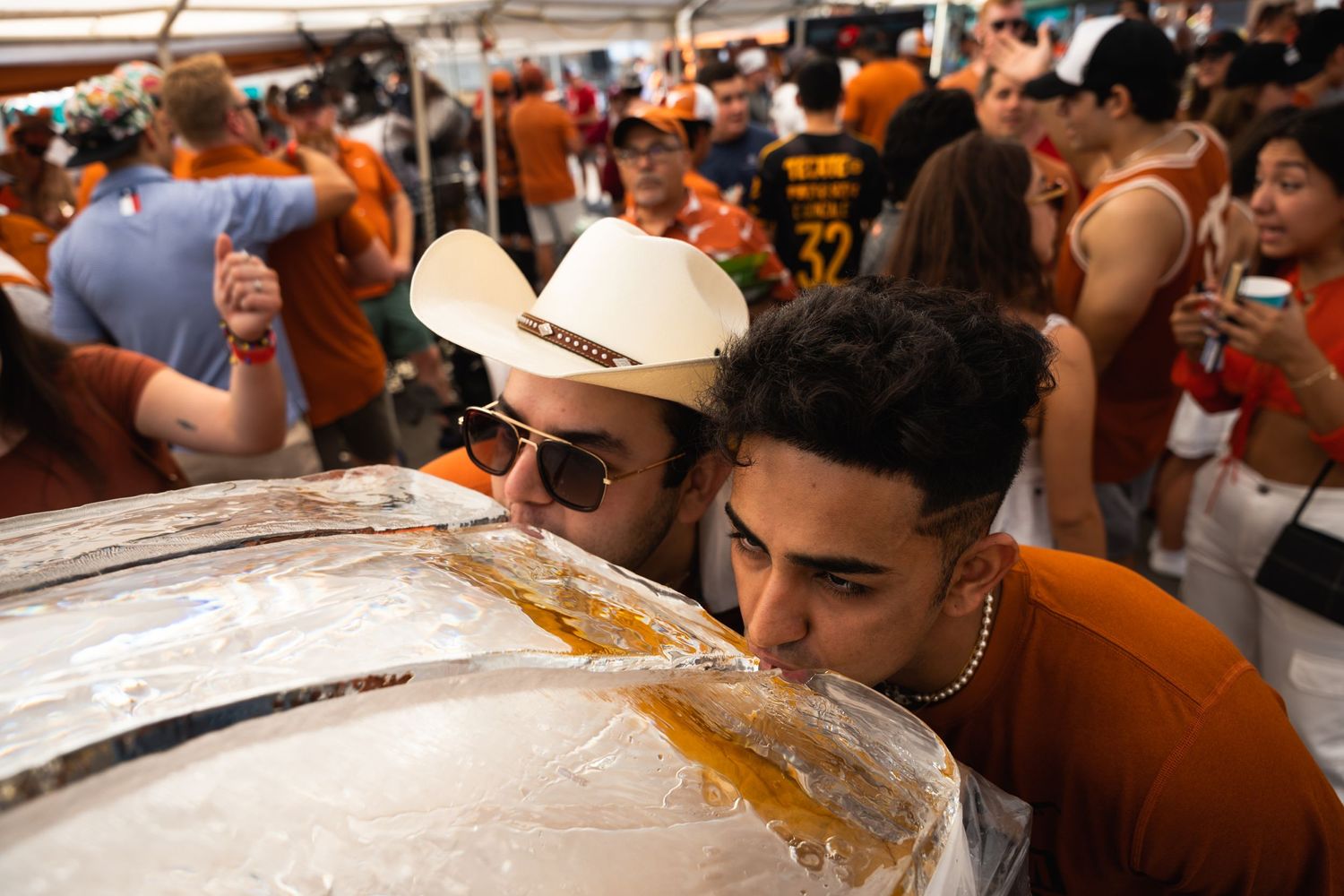 Crowd gathers around ice luge for drinks at outdoor festival with orange-clad attendees.