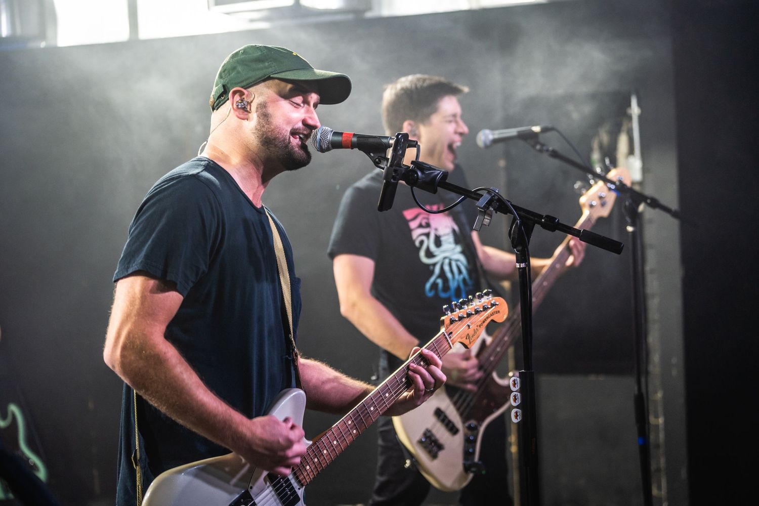 Two musicians perform on stage with guitars while singing into microphones during a live concert.