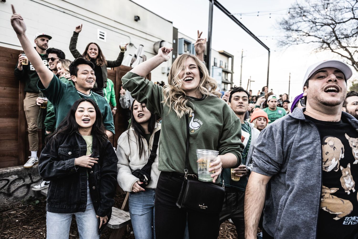 Excited crowd cheers and raises drinks during outdoor event with industrial backdrop.