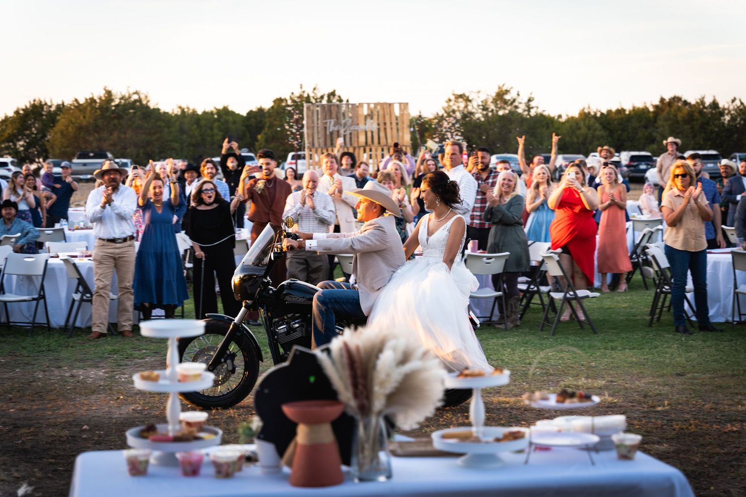 Wedding guests seated outdoors at a rustic ceremony with table settings and decor at sunset.