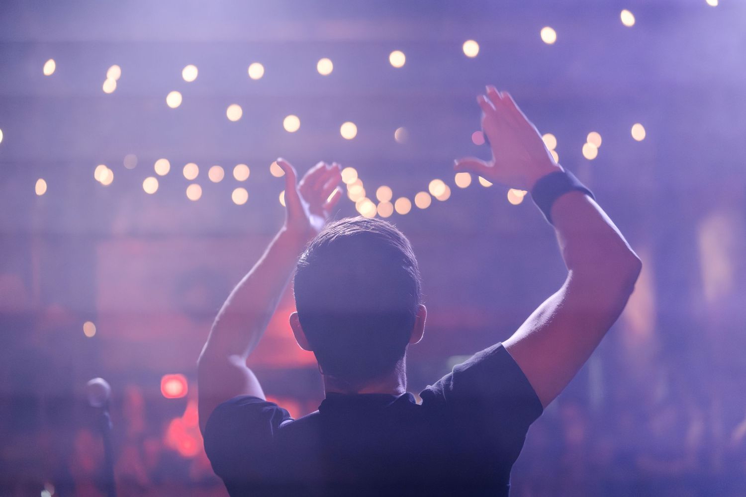 A silhouetted figure holds up hands against a backdrop of string lights at a concert venue.