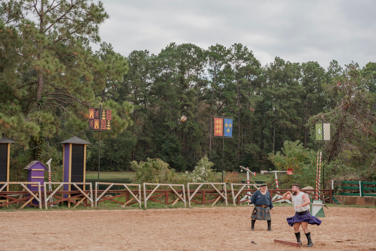 Two people practicing combat moves at renaissance faire with wooden buildings in background.