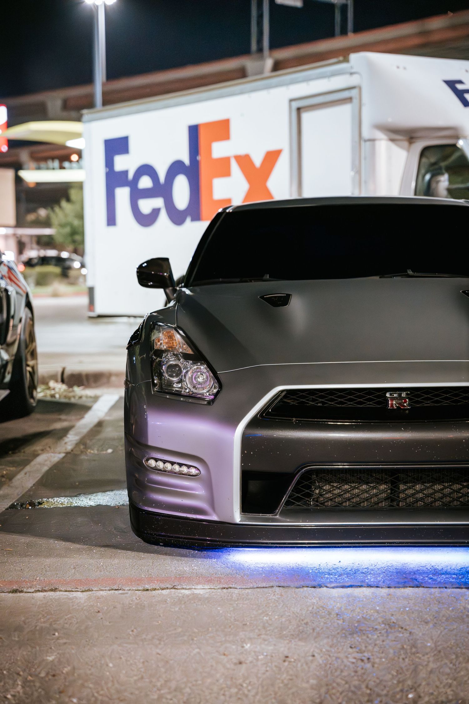 Gray Nissan GT-R with LED underglow parked in front of FedEx sign at night.