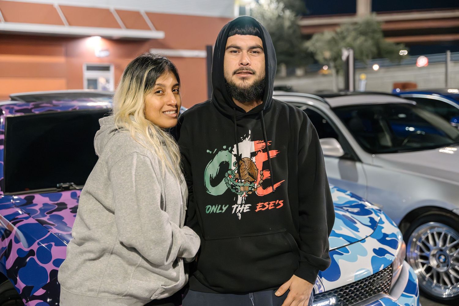 Two people pose in casual clothing at night in a parking lot with custom cars in the background.