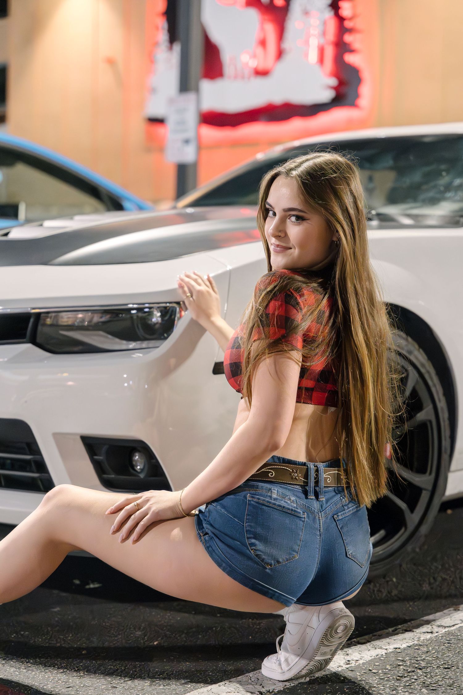 Someone in a red plaid top and denim shorts poses next to a white car.