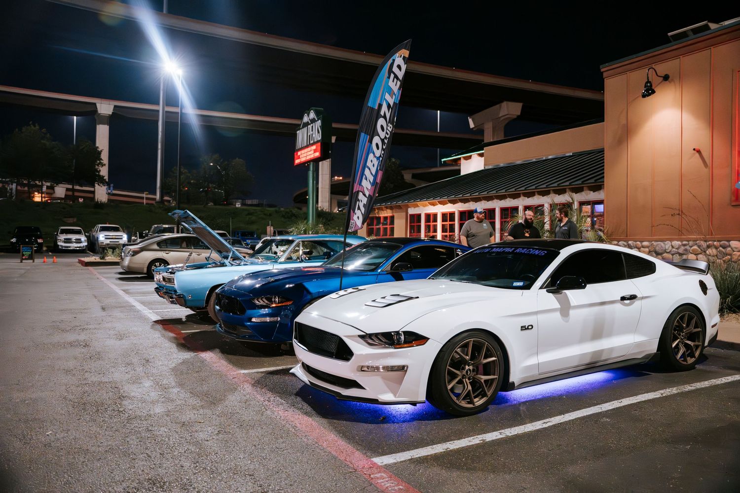 Row of modified sports cars parked at a nighttime car meet with LED lighting.