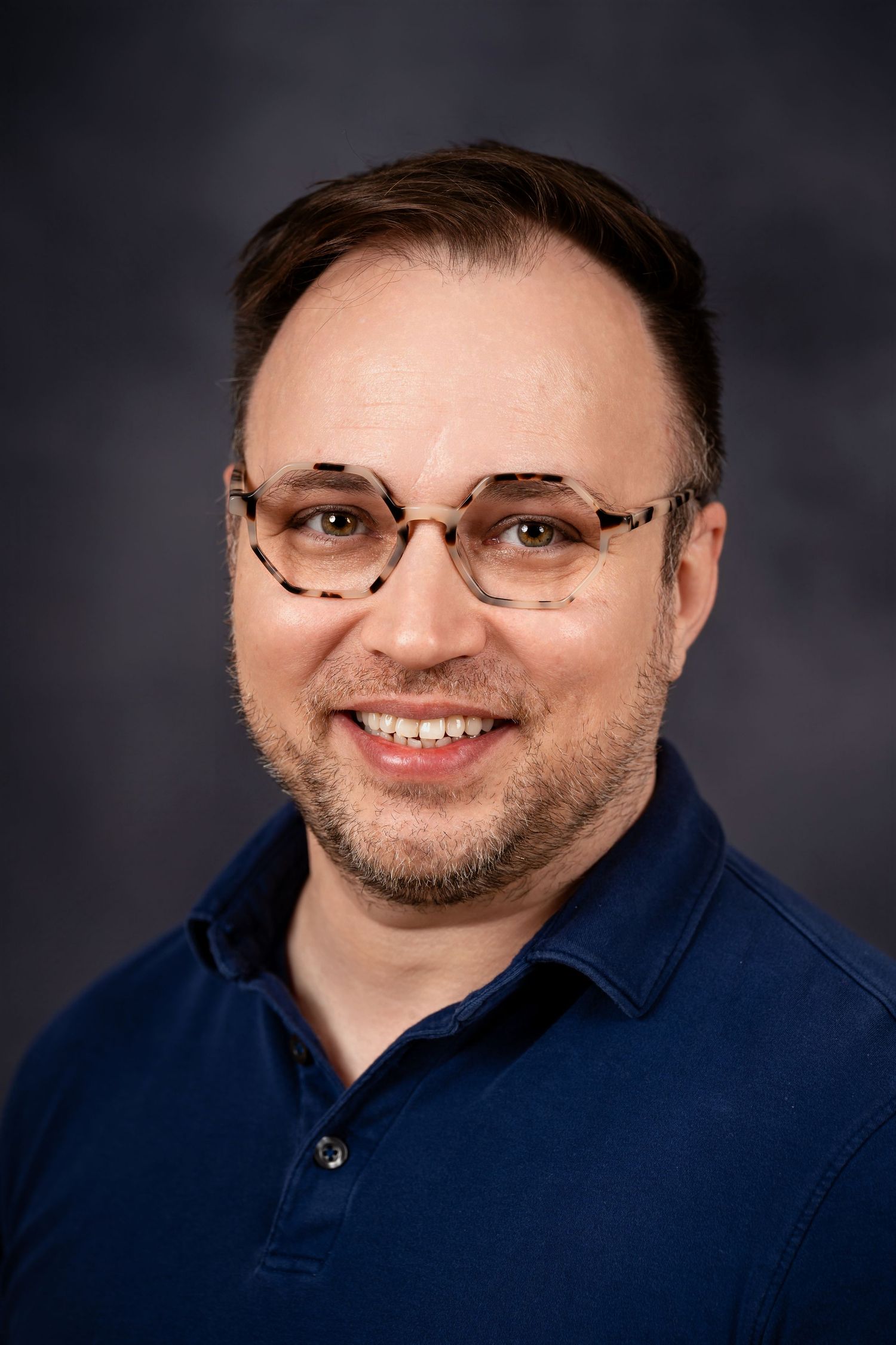 Professional headshot of person in blue polo and glasses against dark background.