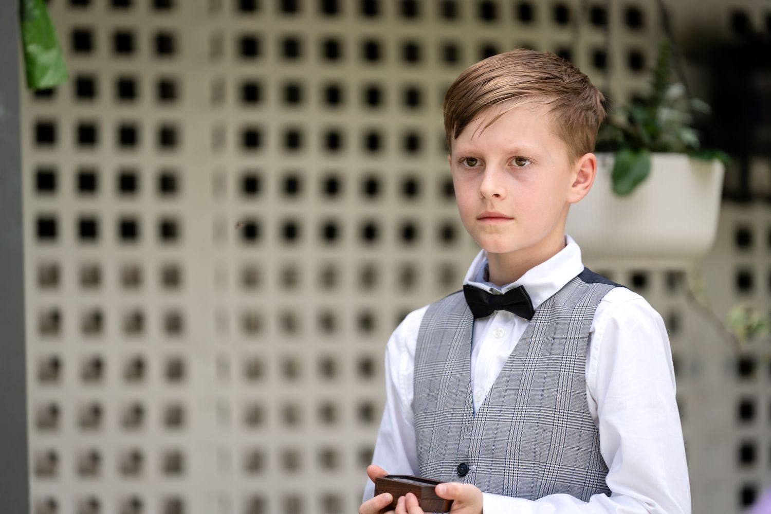 Young attendant in formal gray vest and bowtie poses at an outdoor wedding venue.