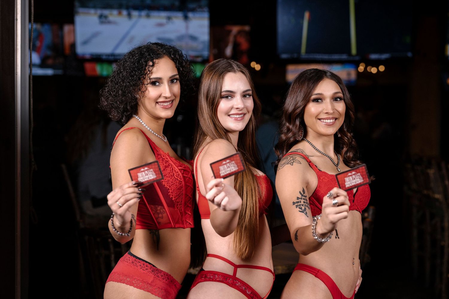 Three women in red lingerie hold cocktails at a bar.