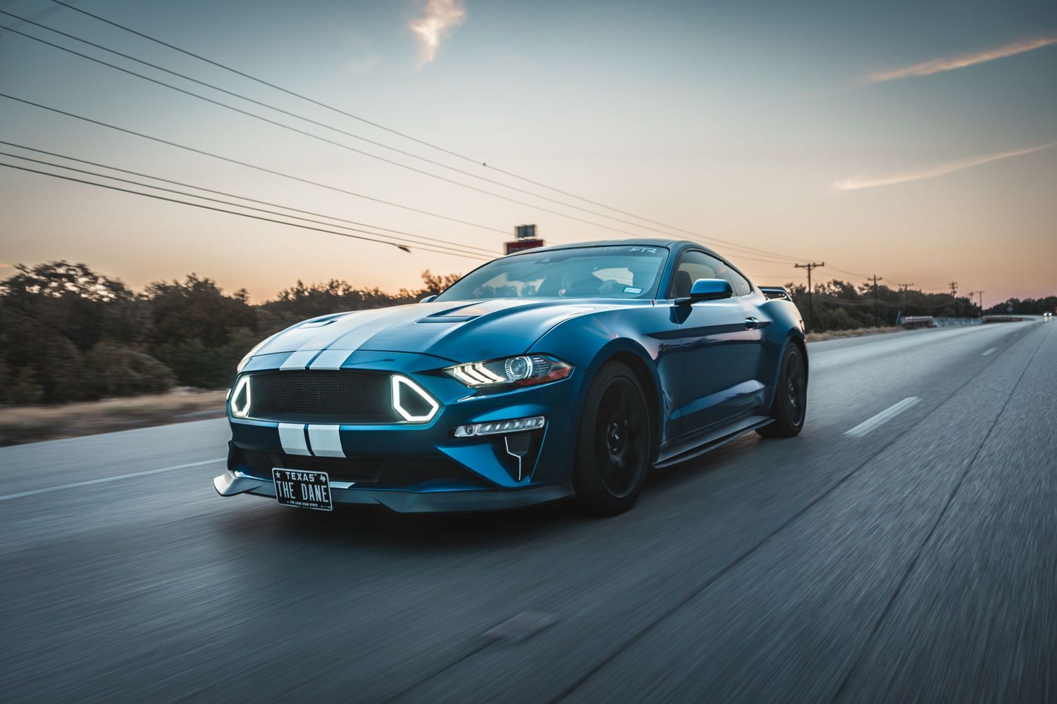 Racing blue Mustang with white stripes captured in motion during sunset.