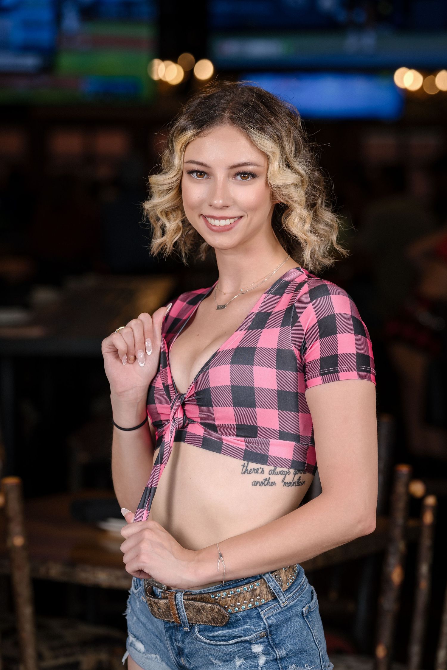 Server in pink plaid tied top and denim poses in restaurant environment.