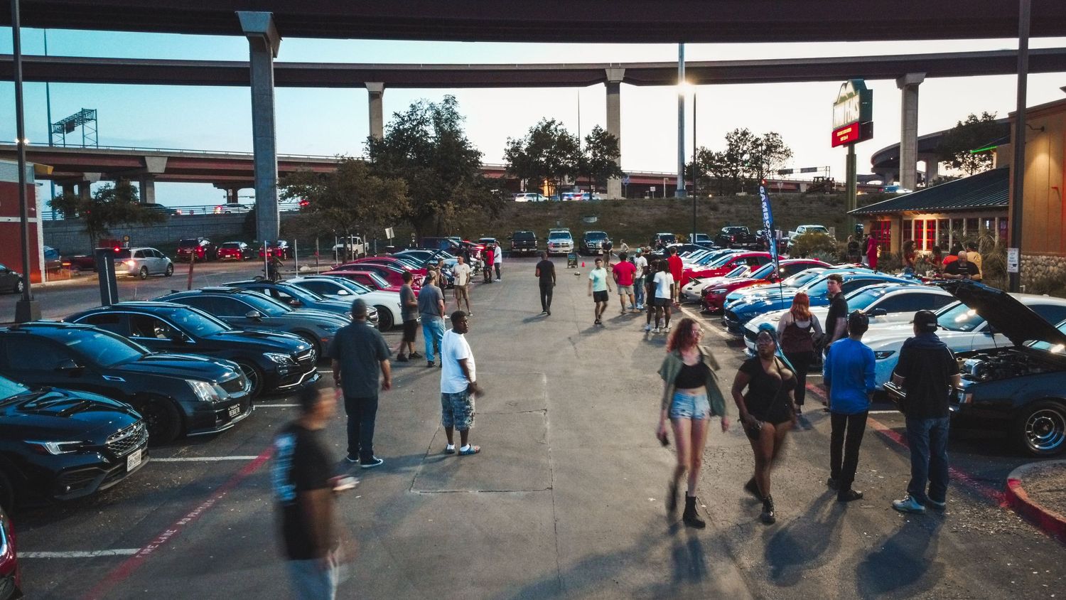 People walking through a parking lot filled with modified cars at an evening car meet event.