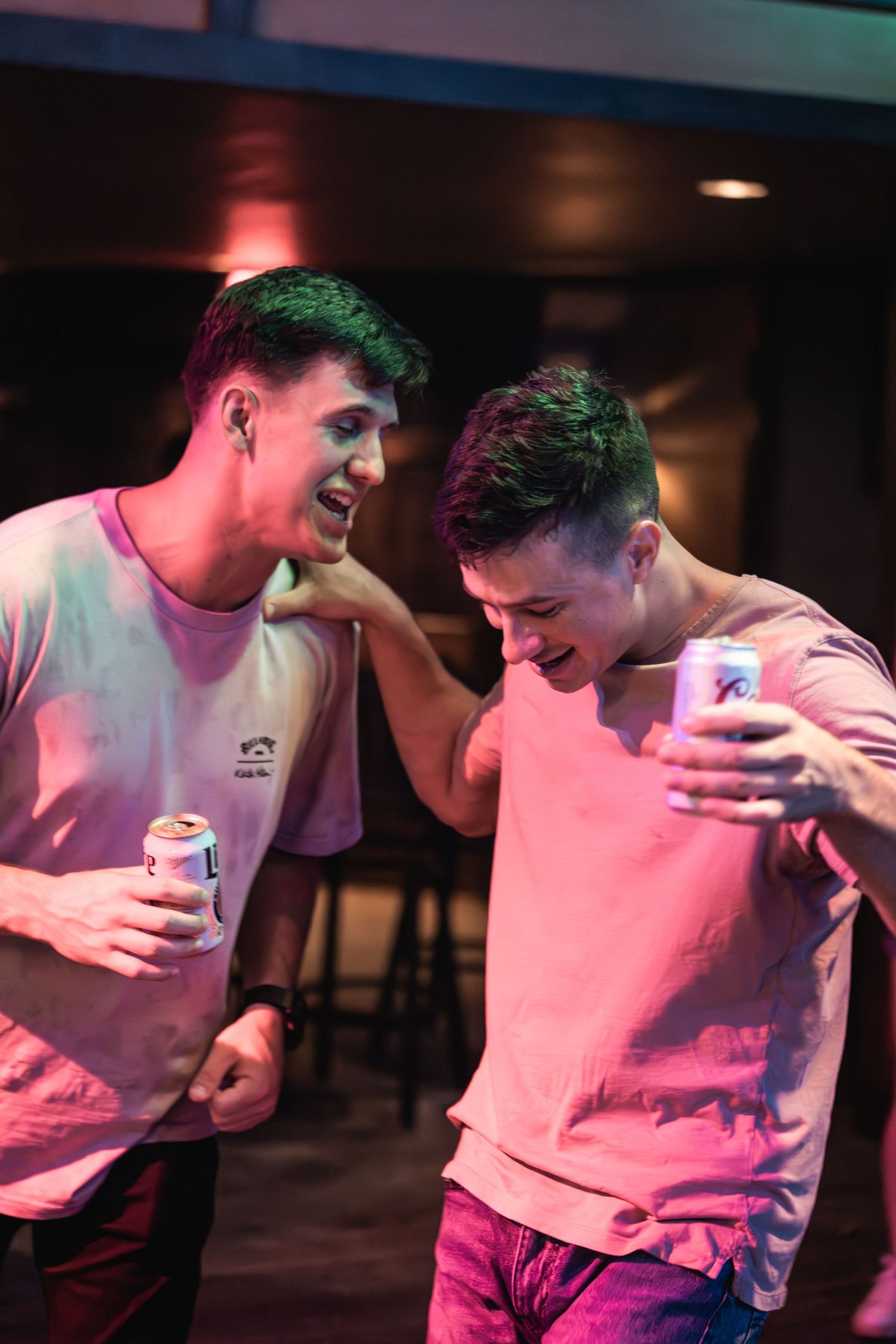 Two friends sharing laughs and drinks at a nightclub under pink lighting.