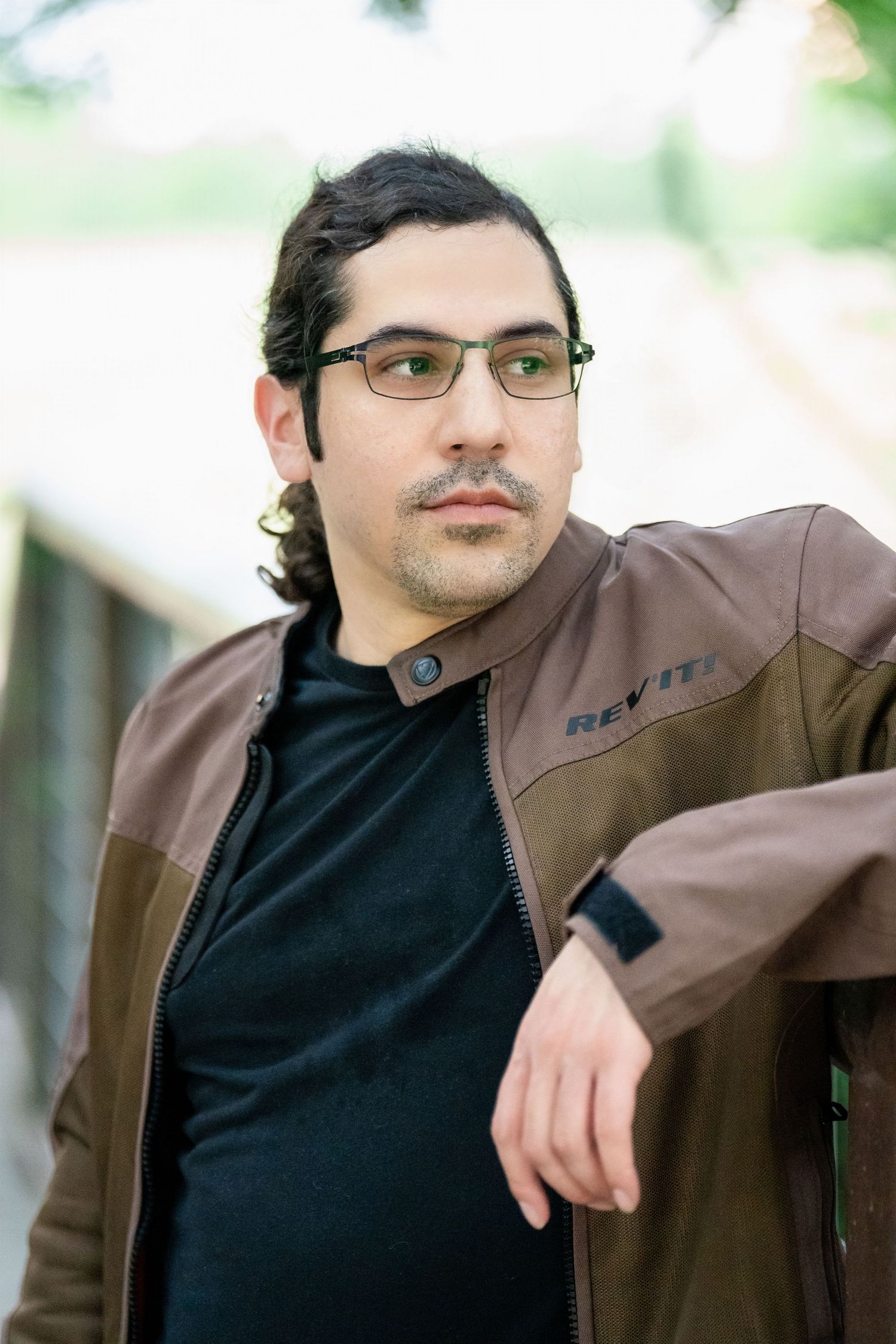 Professional portrait wearing brown leather jacket and black shirt in natural setting.
