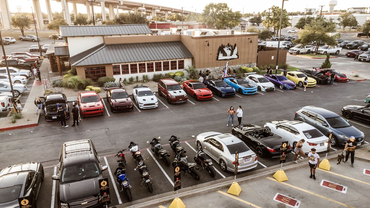 Aerial view of a car meet at A&W restaurant with various modified vehicles in the parking lot.