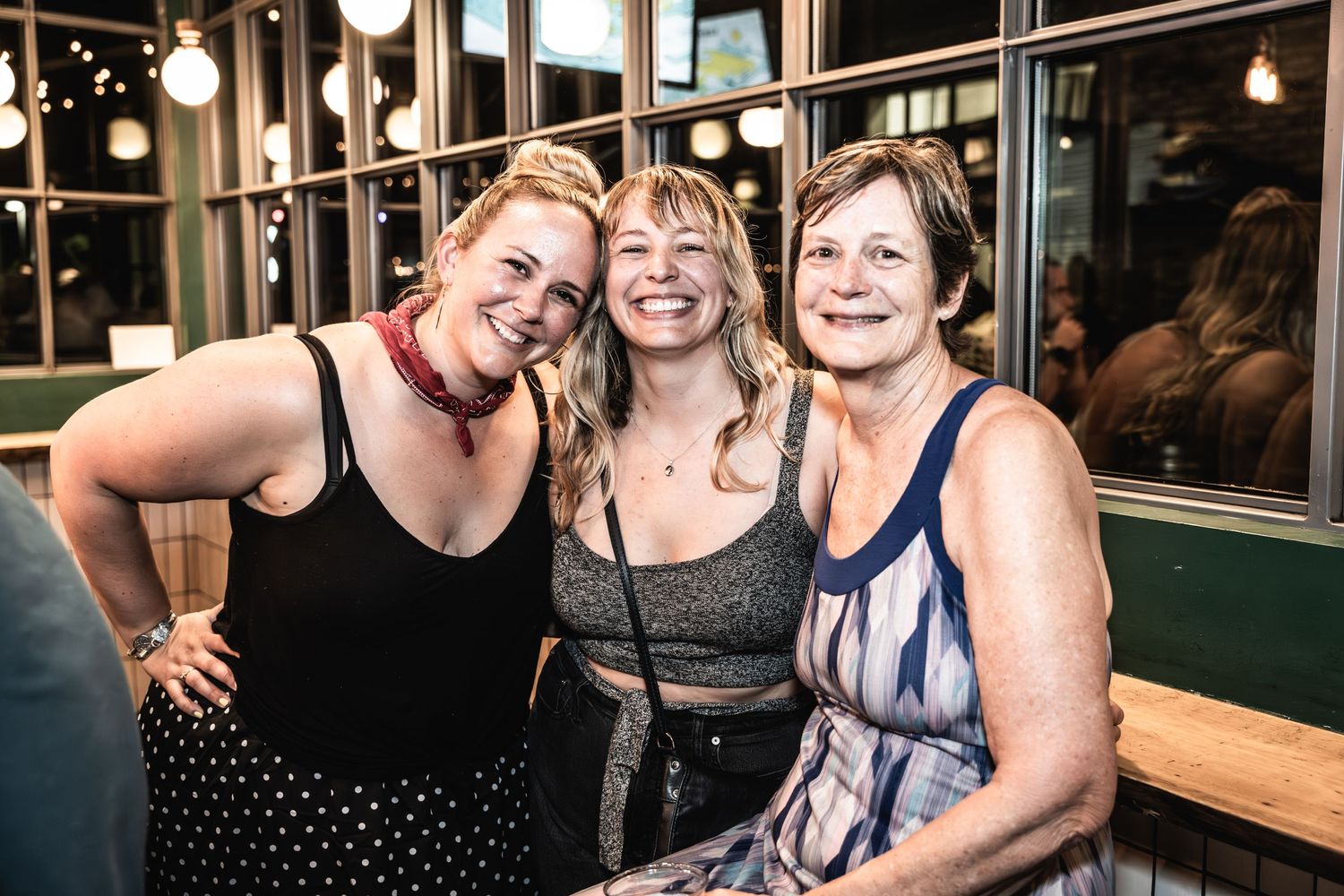 Three friends smiling together at an outdoor venue with windows in the background.