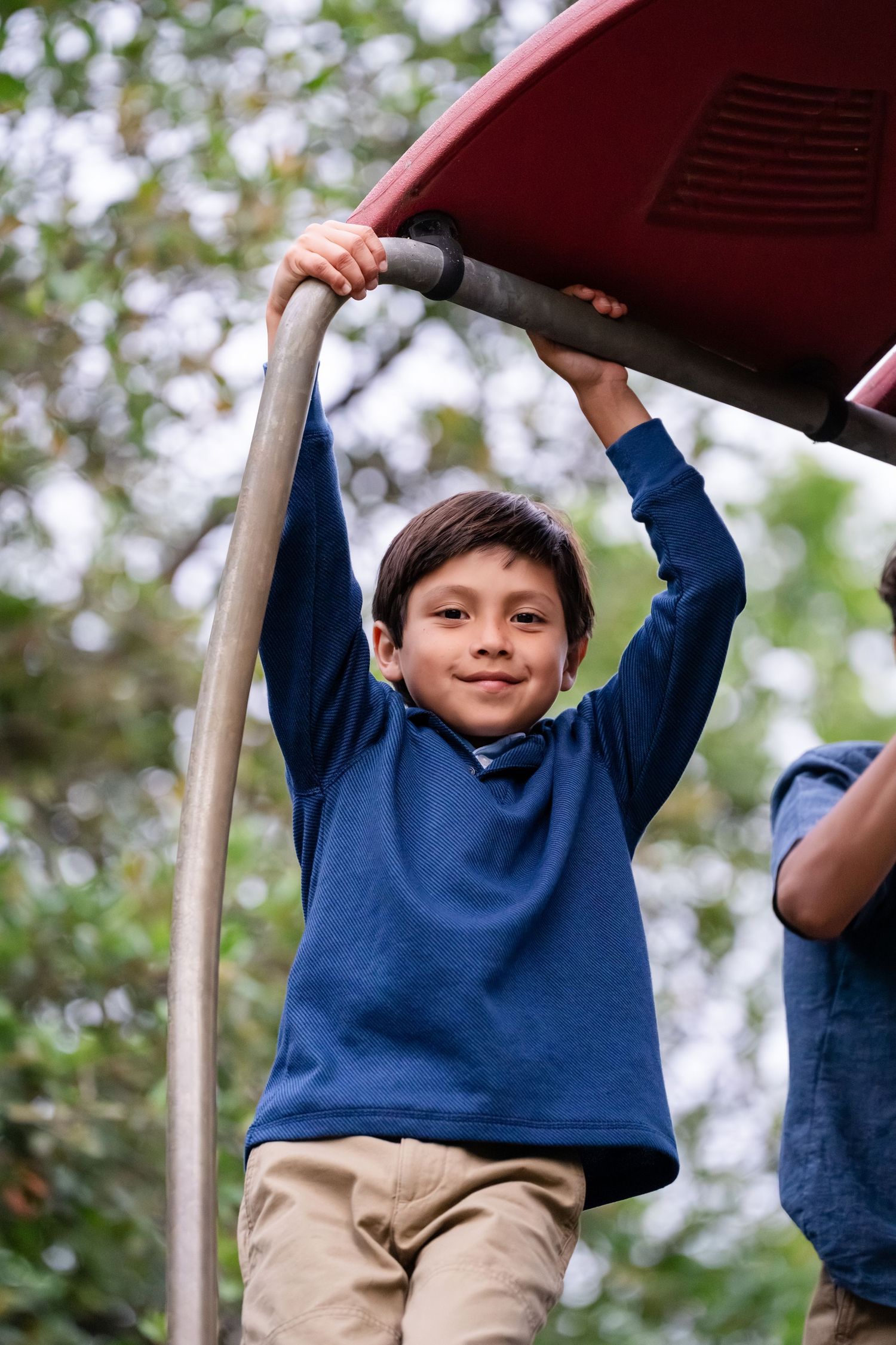 A young boy in a blue sweater smiles while hanging from playground equipment outdoors.