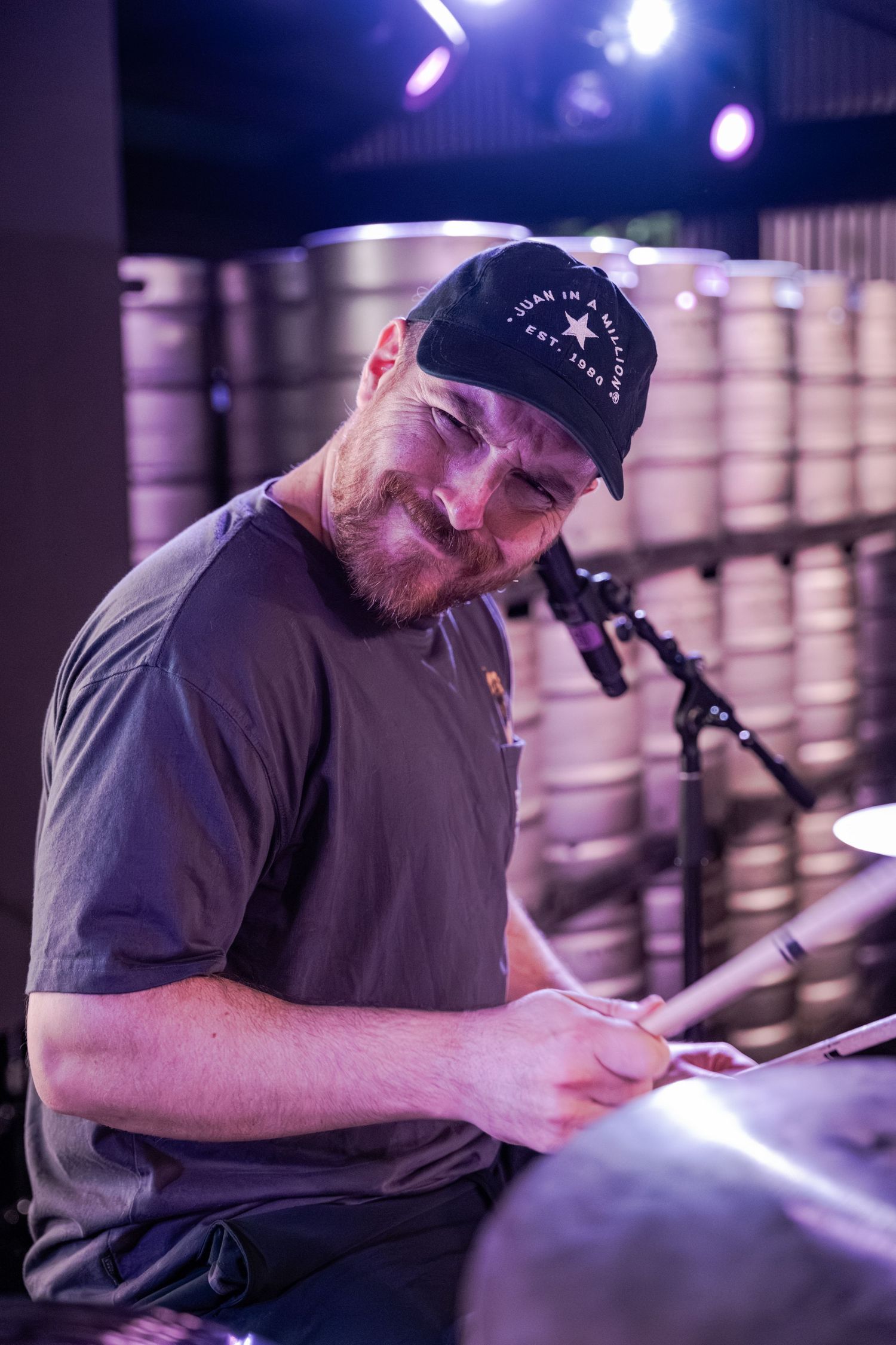 Bearded drummer wearing a black cap plays cymbals on a dimly lit stage with kegs in the background.