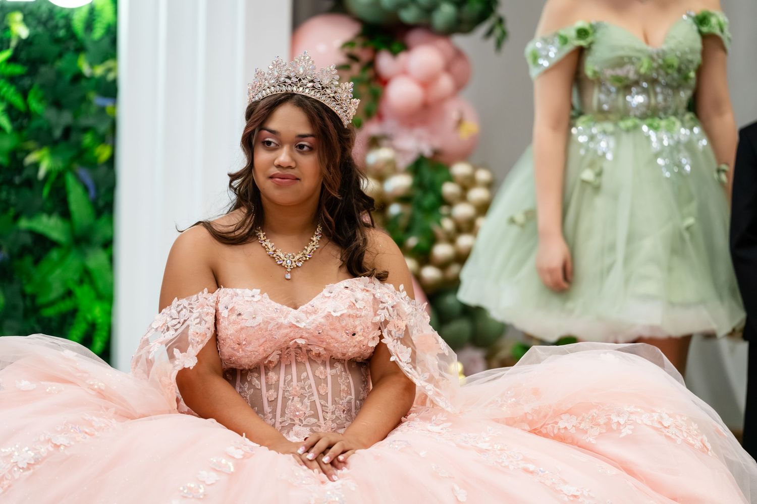 Young woman in pink quinceañera ball gown with tiara and necklace poses at elegant party with balloon decor.