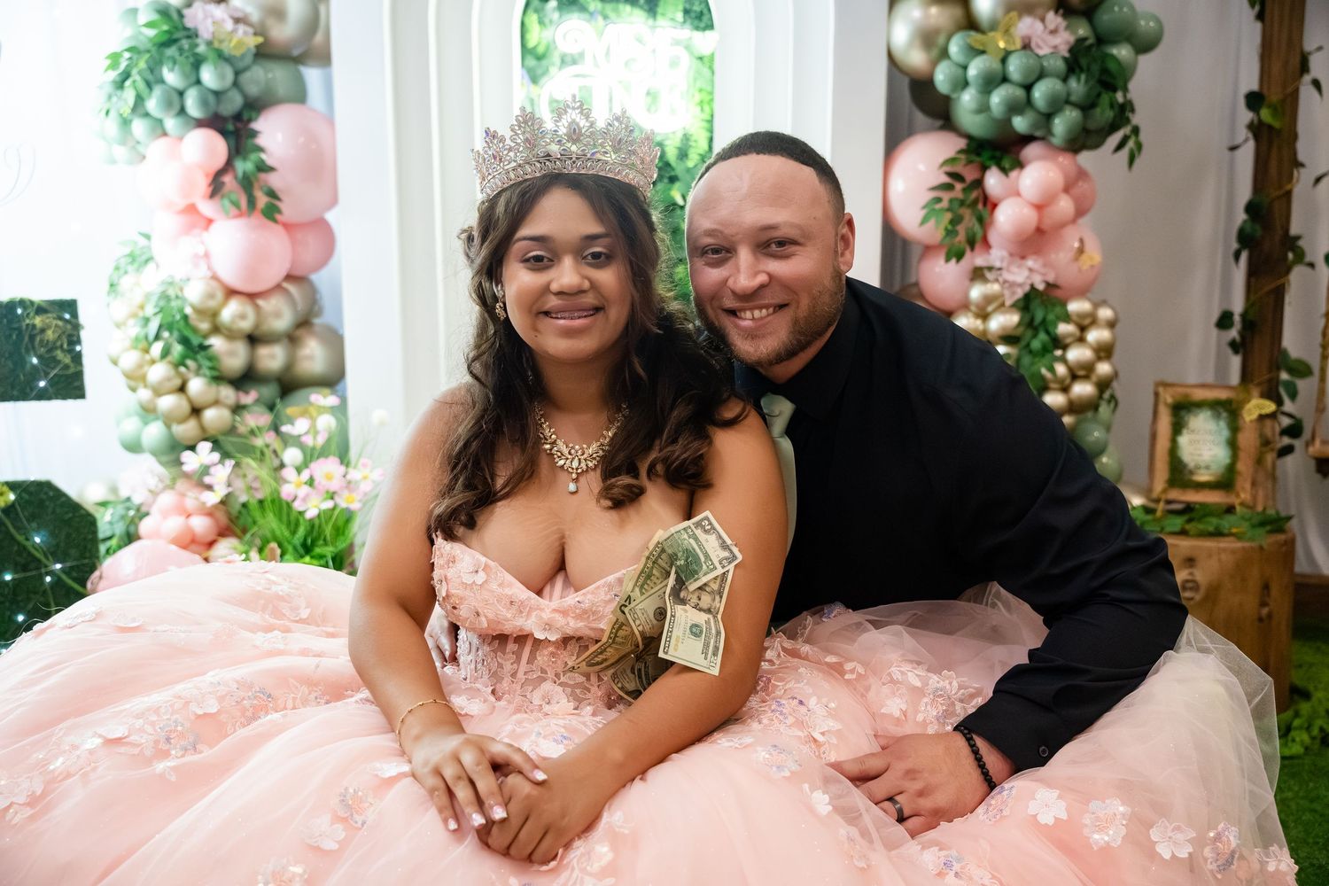 A young woman in a pink quinceañera gown with a tiara poses with a man in a decorated balloon backdrop.