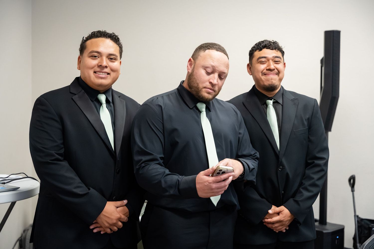 Three men in black suits with green ties pose together, one checking his phone, at a formal event.