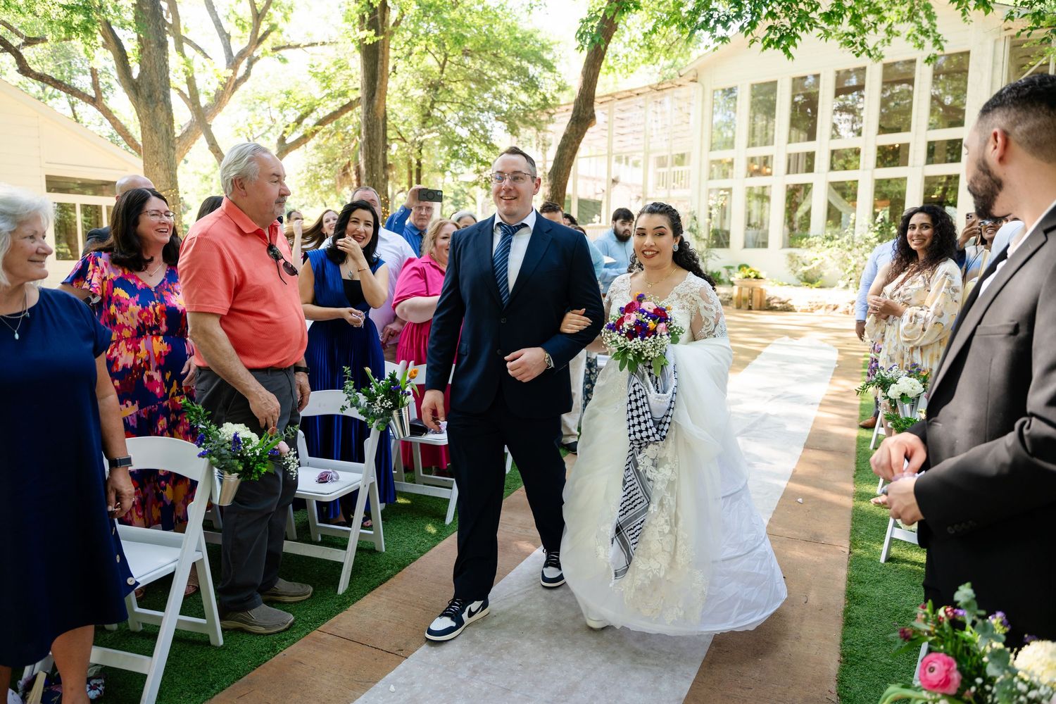 Bride in lace gown walks down outdoor aisle with escort, guests watching, colorful bouquet, sunny garden wedding ceremony.