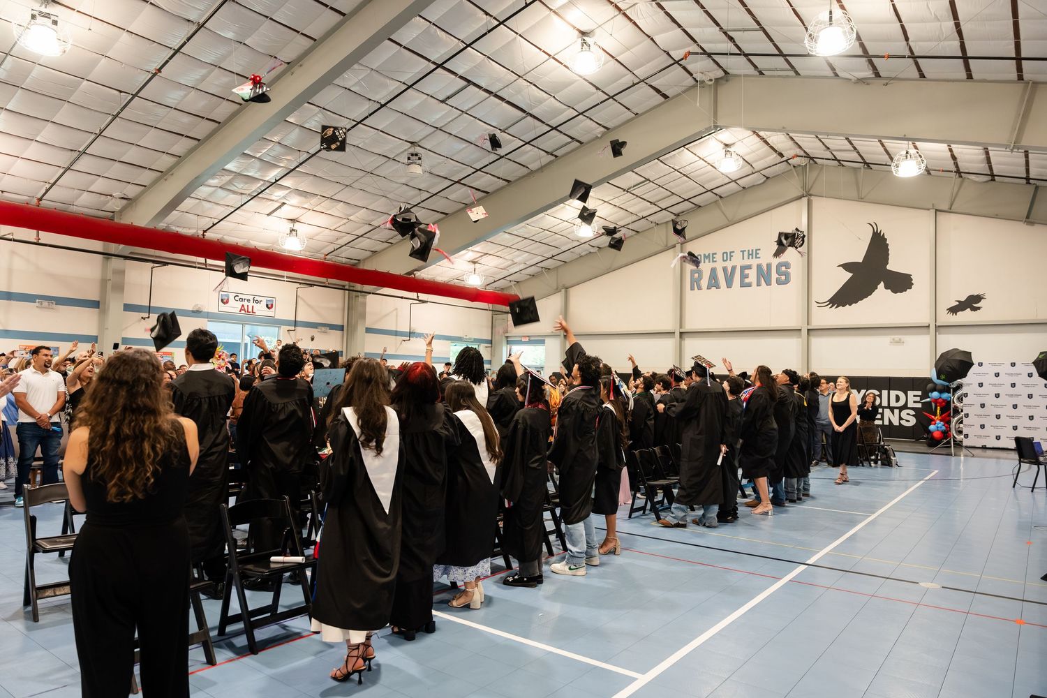 Graduates in black robes toss caps during a Ravens school graduation ceremony held indoors in a decorated gymnasium.